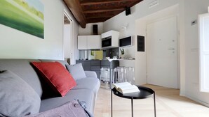 A modern living room with a grey sofa, red pillow, and a small black side table with a book. A kitchen with white cabinets and a tiled backsplash is visible in the background. - Il mulino di Trastevere - G2 (Rome)