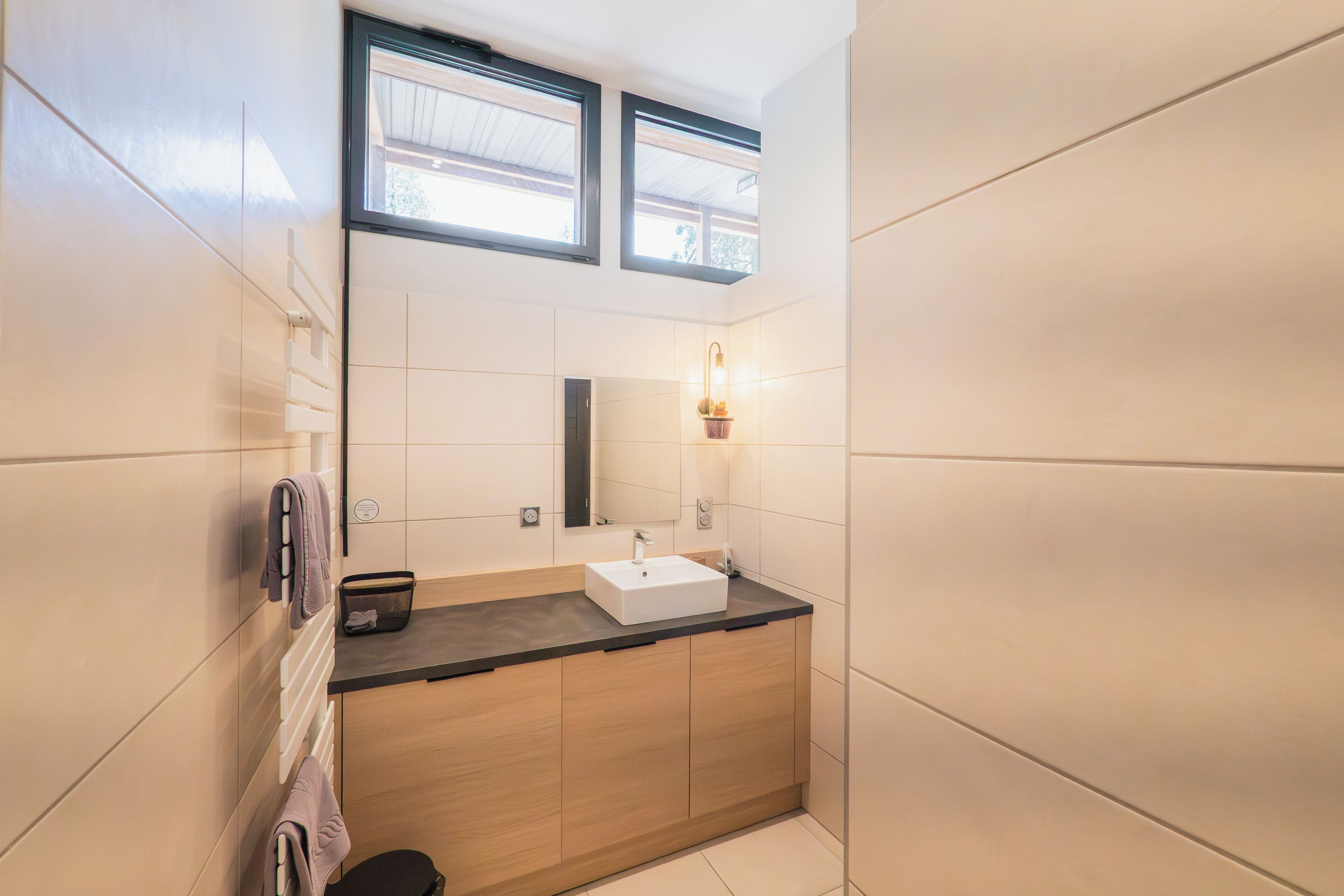 Een moderne badkamer met een houten wastafelmeubel, een witte wasbak en een spiegel. Twee ramen zorgen voor natuurlijk licht.