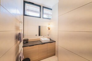 Une salle de bain moderne avec un meuble-lavabo en bois, un lavabo blanc et un miroir. Deux fenêtres laissent entrer la lumière naturelle.