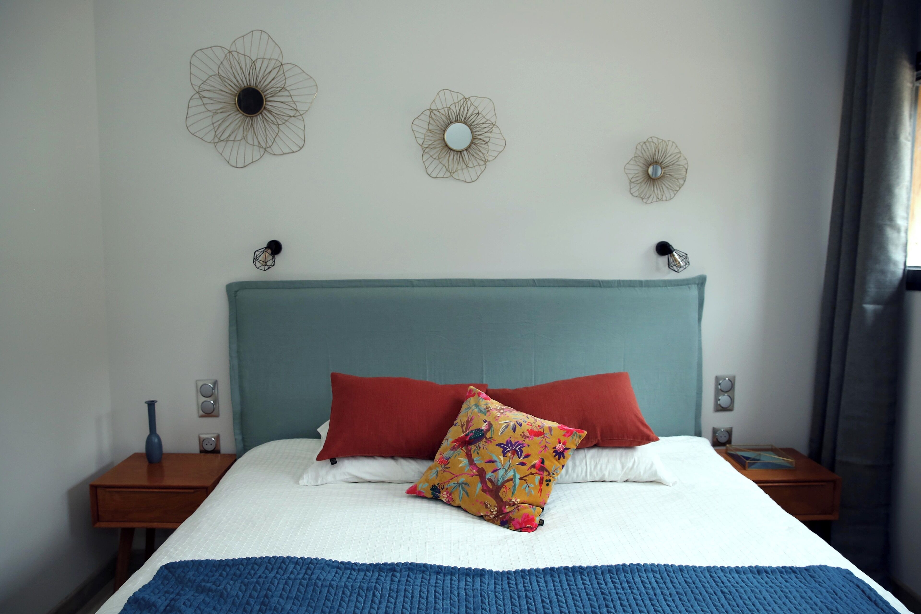 Una cama bien hecha con un edredón azul, dos almohadas rojas y una almohada con motivos florales de colores. Hay tres lámparas fijadas a la pared y una mesita de noche con un florero a la izquierda.