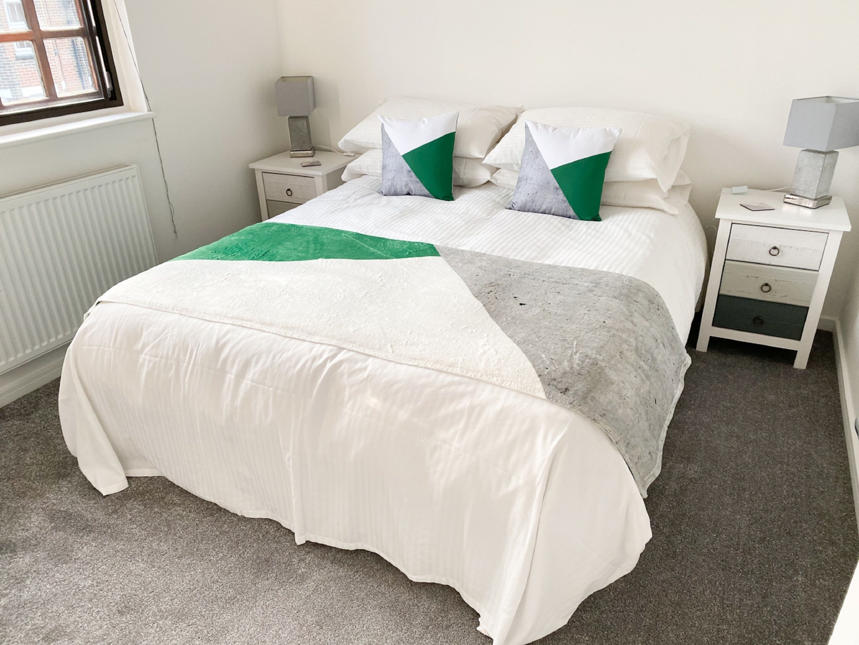 Una cama bien hecha con una colcha blanca y verde, dos mesitas de noche con lámparas y una ventana con persianas.