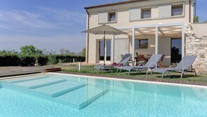 Une piscine avec des chaises longues et un parasol, un mur en pierre et une maison à deux étages dotée de grandes fenêtres.