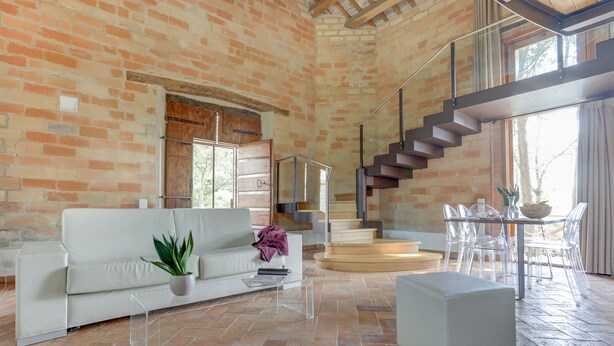 A spacious living area with a brick wall, a white sofa, a glass coffee table, a grey ottoman, and a staircase leading to an upper level.