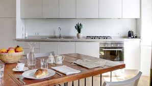 A modern kitchen with white cabinets, a wooden dining table, and a breakfast setup including fruits, a croissant, and a newspaper.