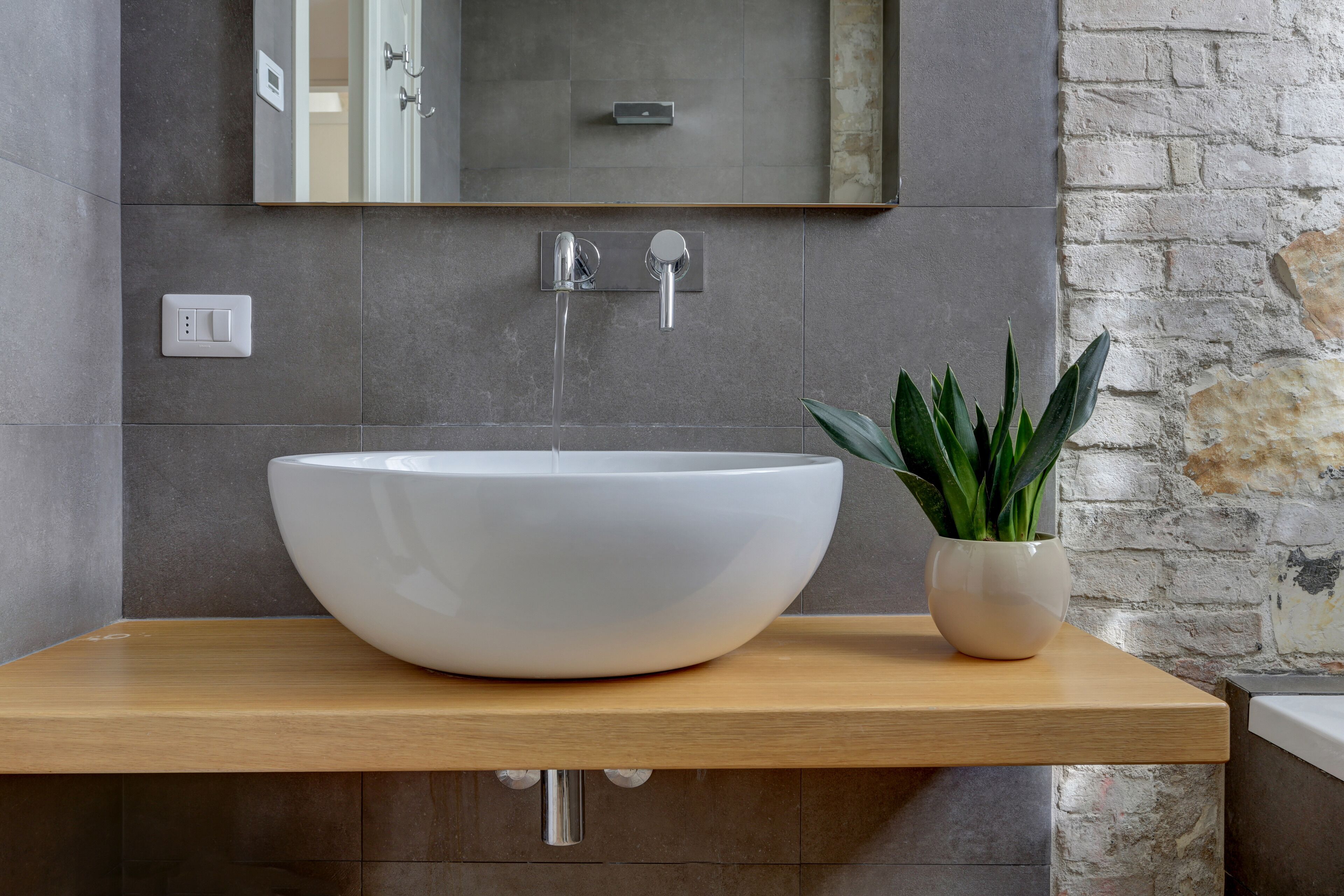 A modern bathroom with a large white vessel sink, a wooden countertop, a potted plant, and a stone wall.