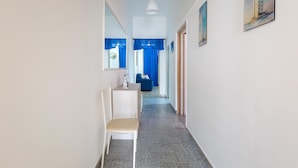 En smal hall med en vit stol, ett litet bord och en spegel på vänster sida. Hallen leder in till ett rum med en blå gardin och en säng.