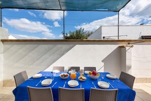 Un área de comedor al aire libre con una mesa preparada para cuatro, cubierta con un mantel azul, rodeada de sillas de mimbre y una pared blanca al fondo.