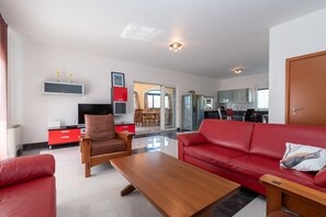 Uma sala de estar moderna com um sofá vermelho, uma poltrona de couro marrom e uma mesa de centro de madeira. Há uma TV de tela plana em um armário vermelho e uma cozinha visível ao fundo.