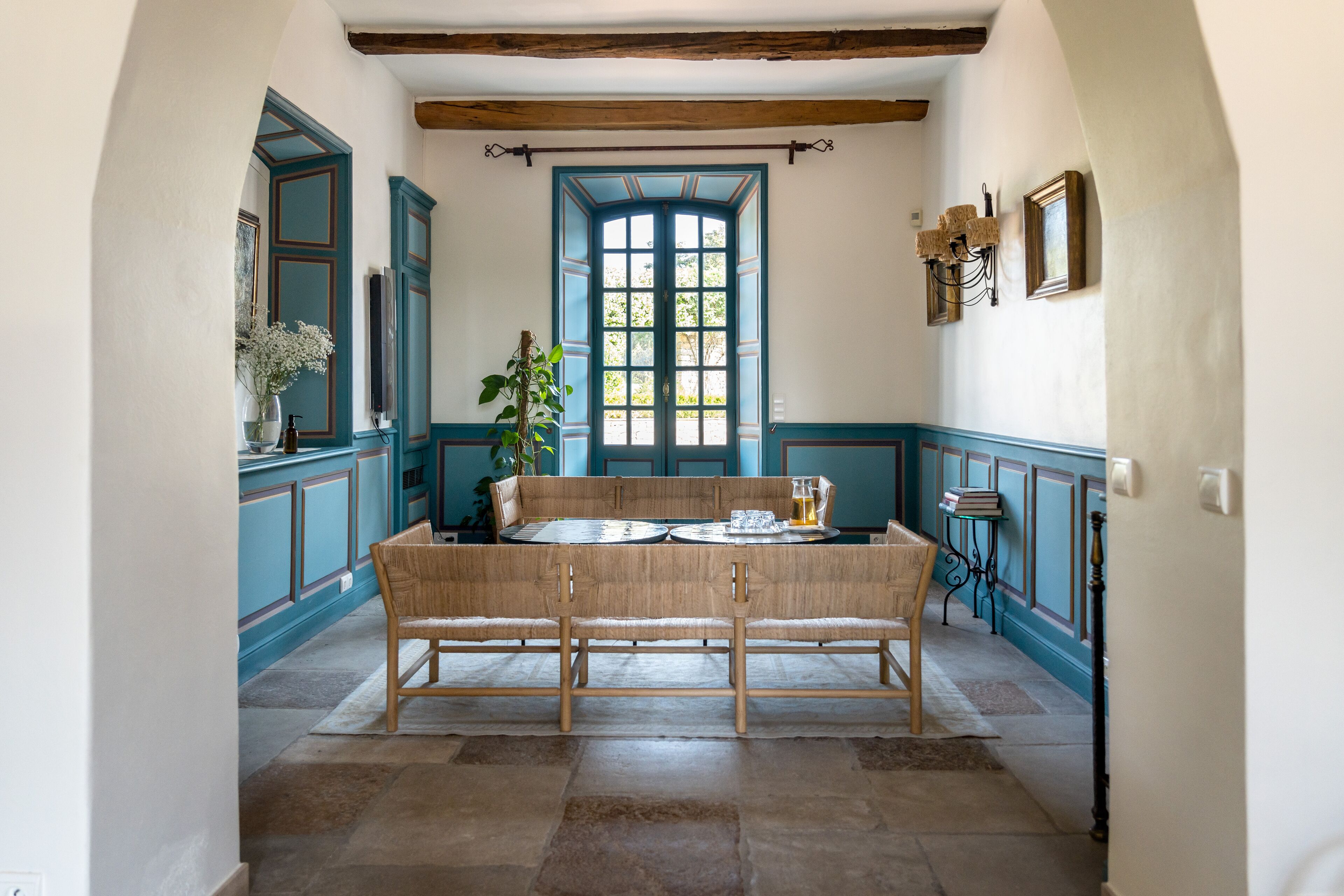 A dining area with a wooden bench and table, a large window with blue shutters, and a potted plant.