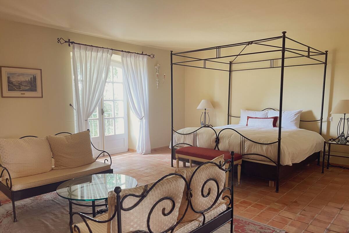 A four-poster bed with a canopy, a sofa, a glass-top table, and a chair in a room with tiled flooring and a large window with curtains.