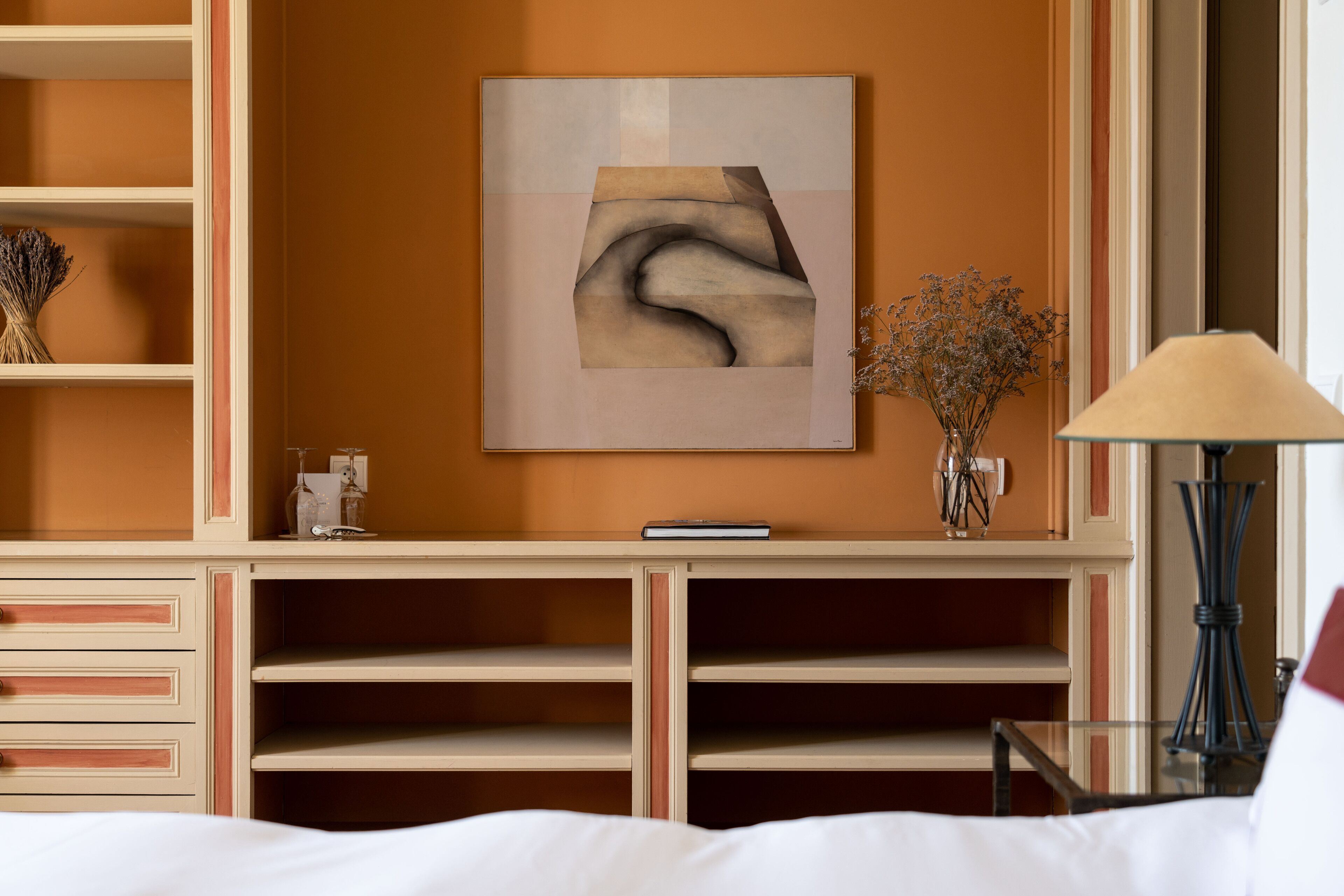 Een hotelkamer met een bed, nachtkastje, lamp, een aan de muur hangend kunstwerk en een vaas met gedroogde bloemen.