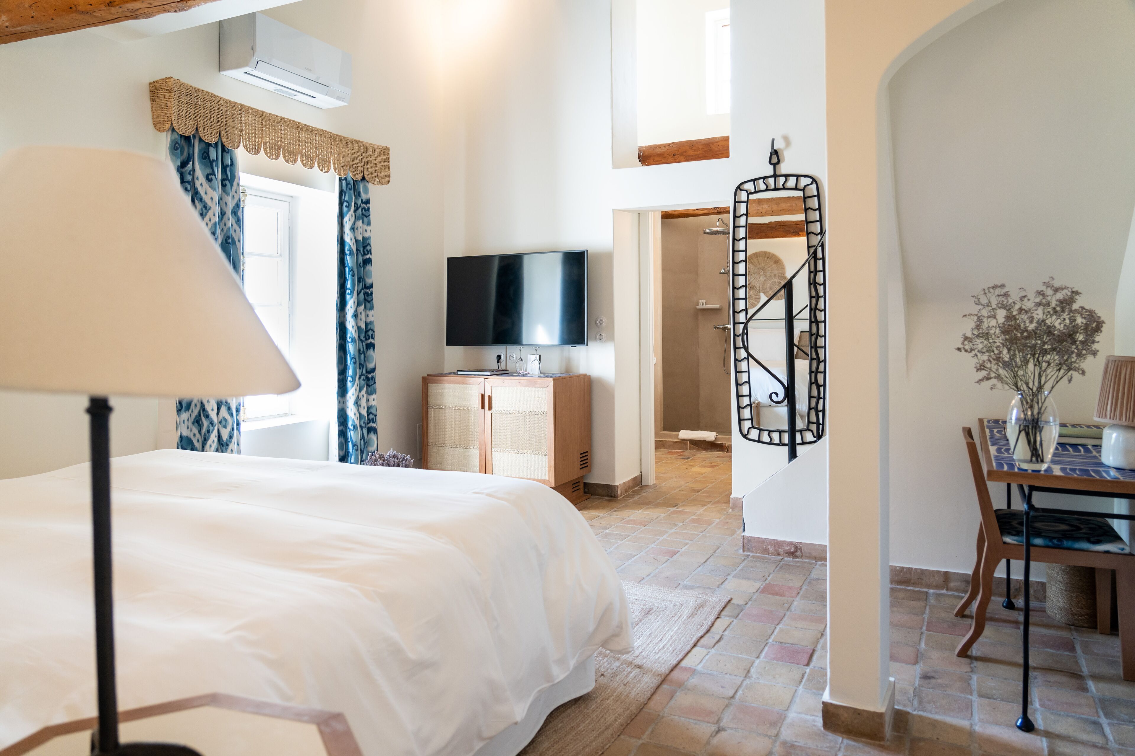 A bedroom with a bed, a television, a desk with a chair, and a shower area.
