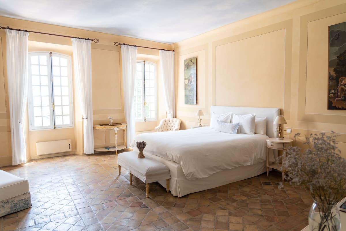A spacious bedroom with a large bed, a bedside table, a chair, and a window with curtains.