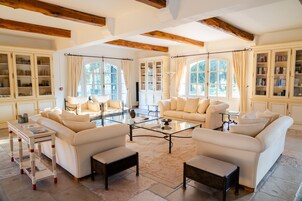 A spacious living room with high ceilings, exposed wooden beams, and large windows. There are multiple seating areas with white sofas and armchairs, a glass coffee table, and built-in bookcases along the walls.