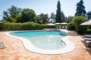 Una piscina con borde ondulado, rodeada por una terraza con baldosas y sillones de descanso con sombrillas.