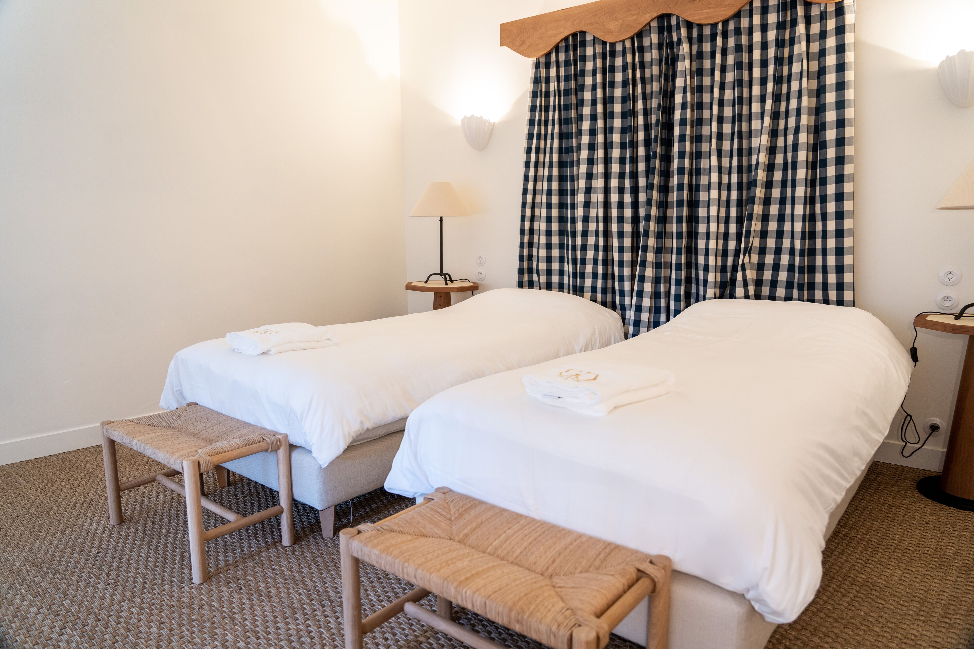 A hotel room with two beds, a small table, a bench, and a lamp.