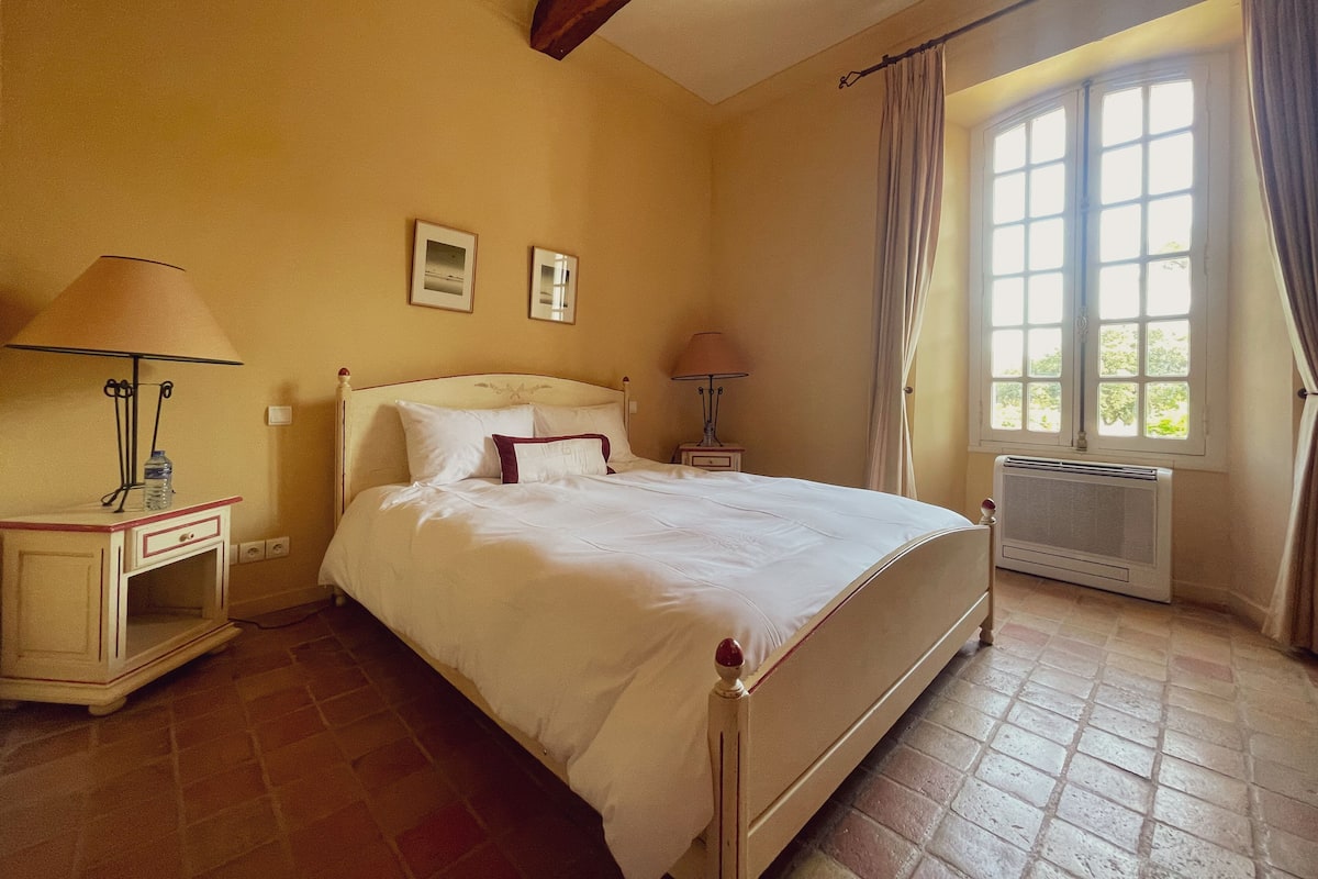 A bedroom with a bed, two bedside tables, a nightstand, a window with curtains, and an air conditioning unit.