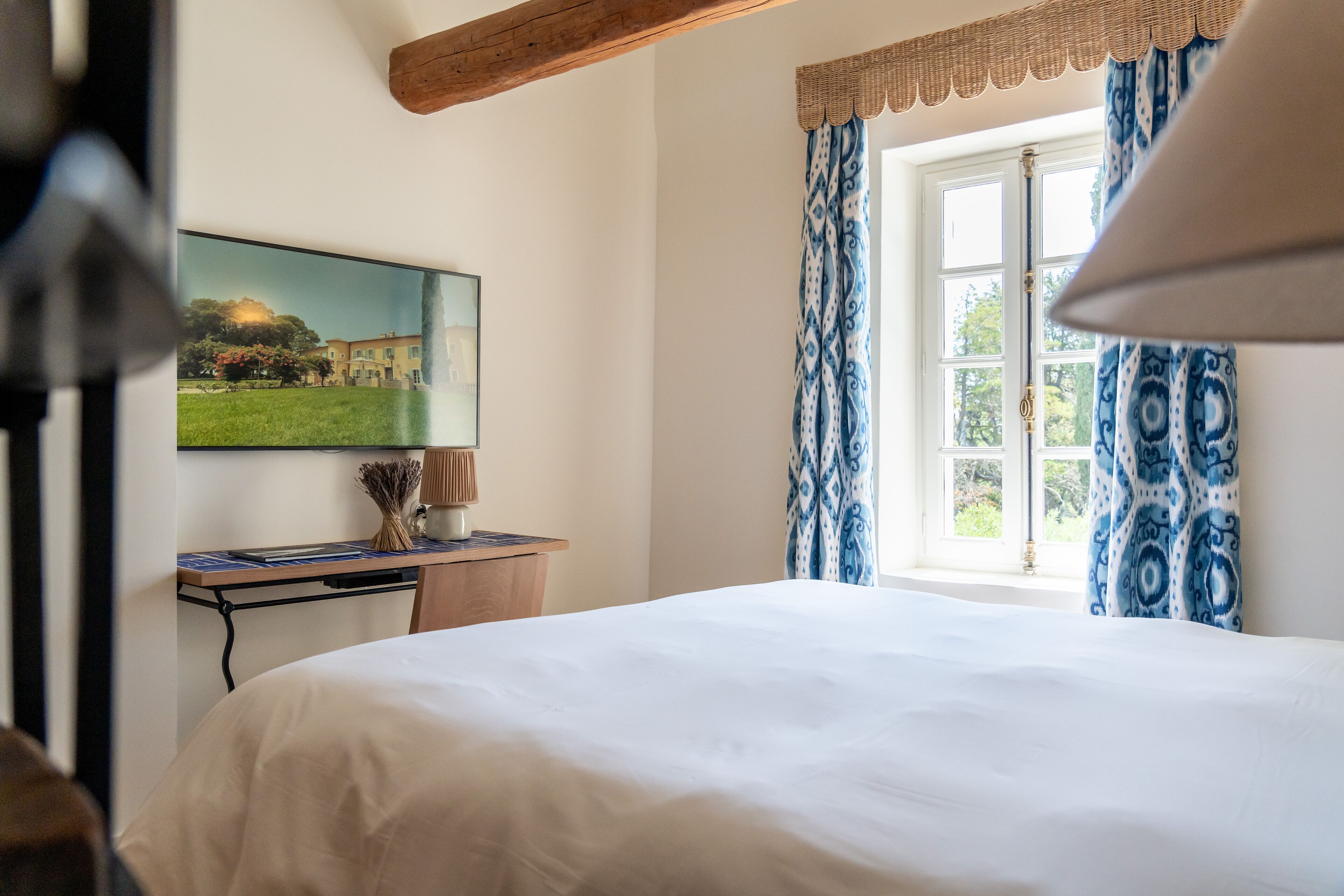A bedroom with a bed, a desk with a lamp, a painting, and patterned curtains.