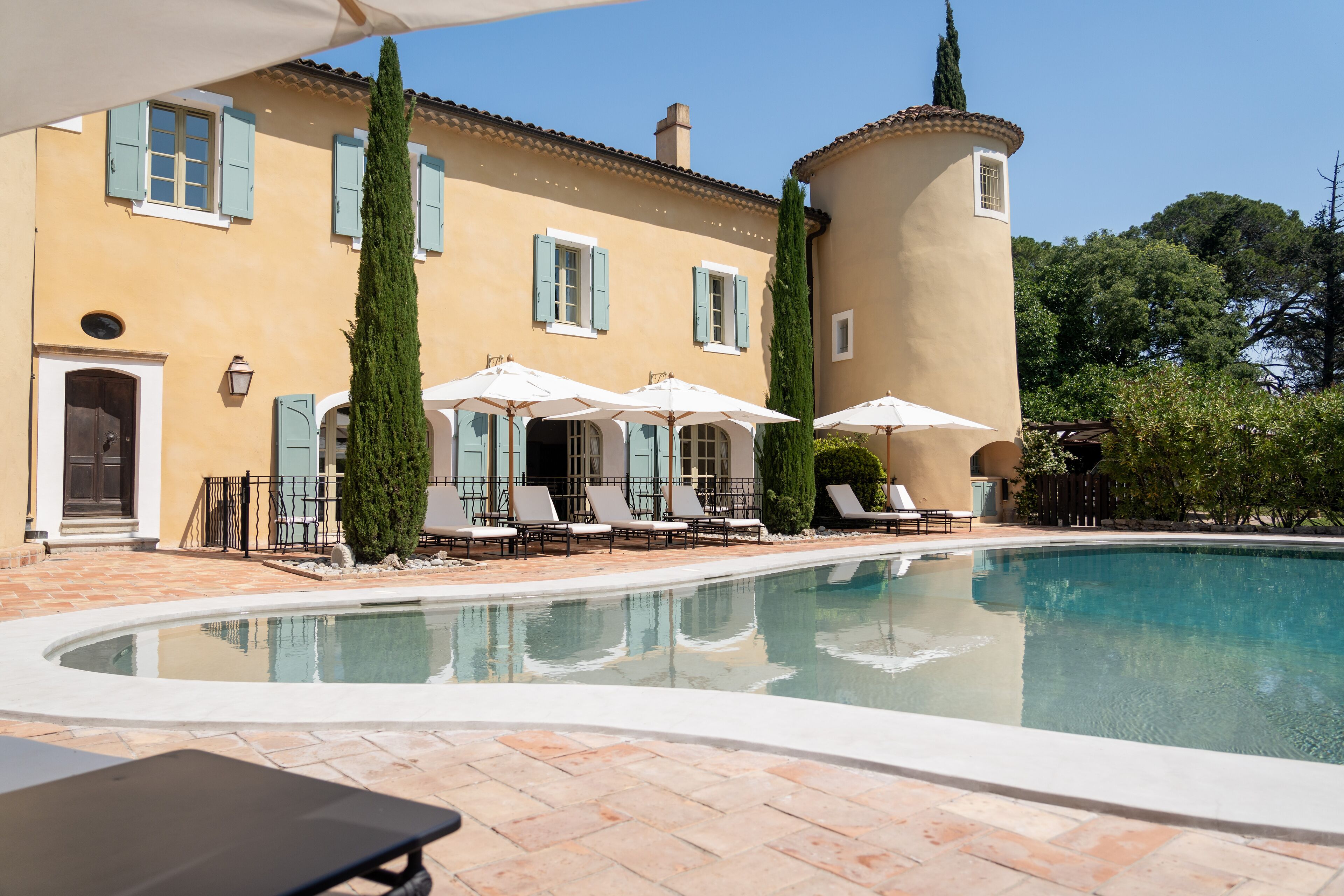 Un espace aménagé au bord de la piscine, avec des chaises longues et des parasols, un bâtiment aux volets bleus et une piscine à l’eau d’un bleu limpide.