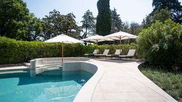 Una piscina de borde curvo, rodeada de sillas de descanso y sombrillas, con árboles y follaje al fondo.