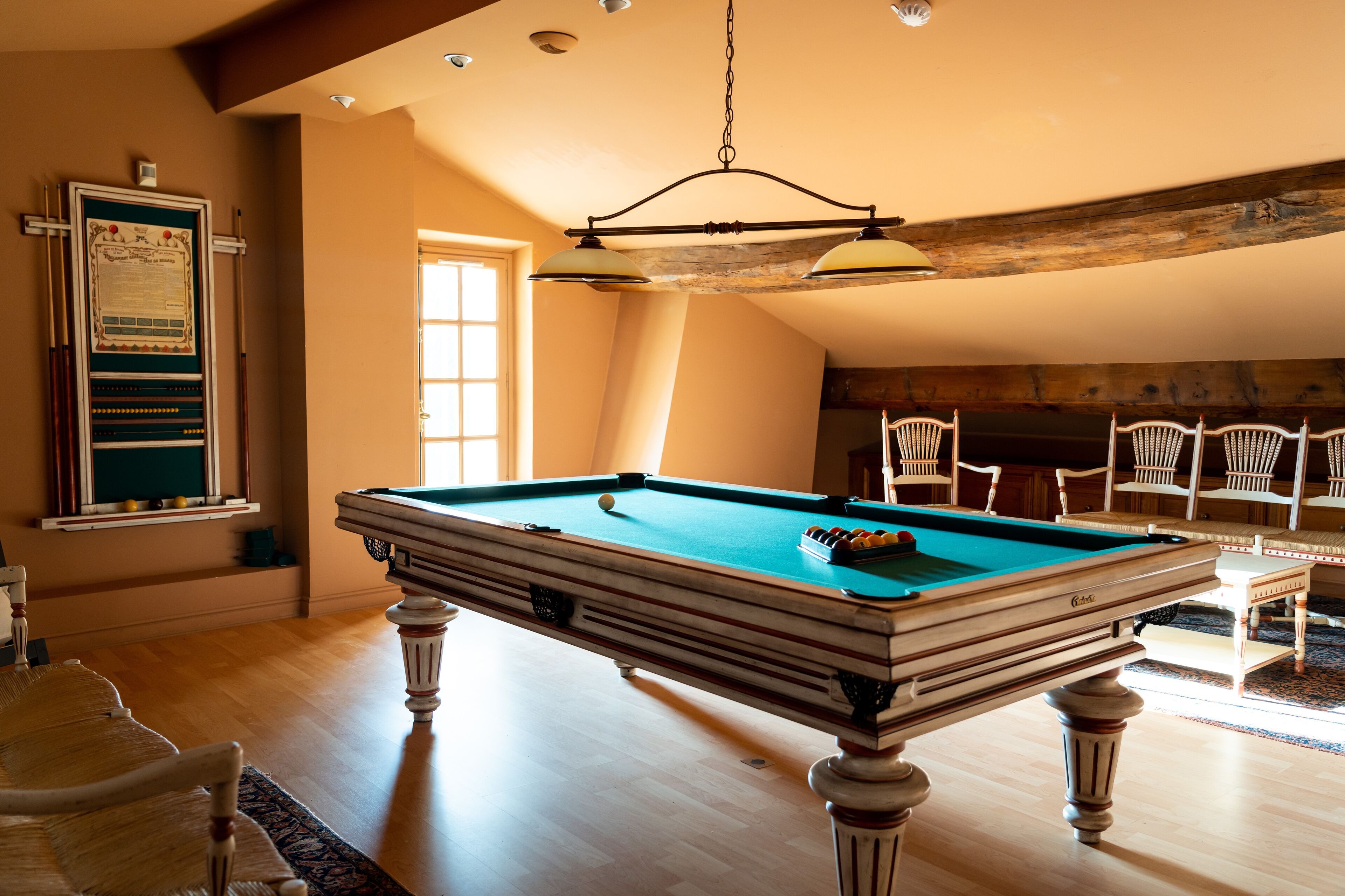 Une table de billard dans une pièce au plancher en bois, avec un plafond haut aux poutres apparentes et plusieurs fauteuils en osier.