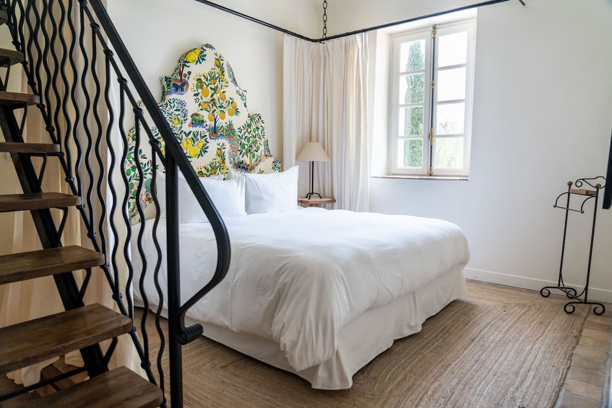 A bedroom with a bed, a wooden staircase, a painting, a window with curtains, and a nightstand.