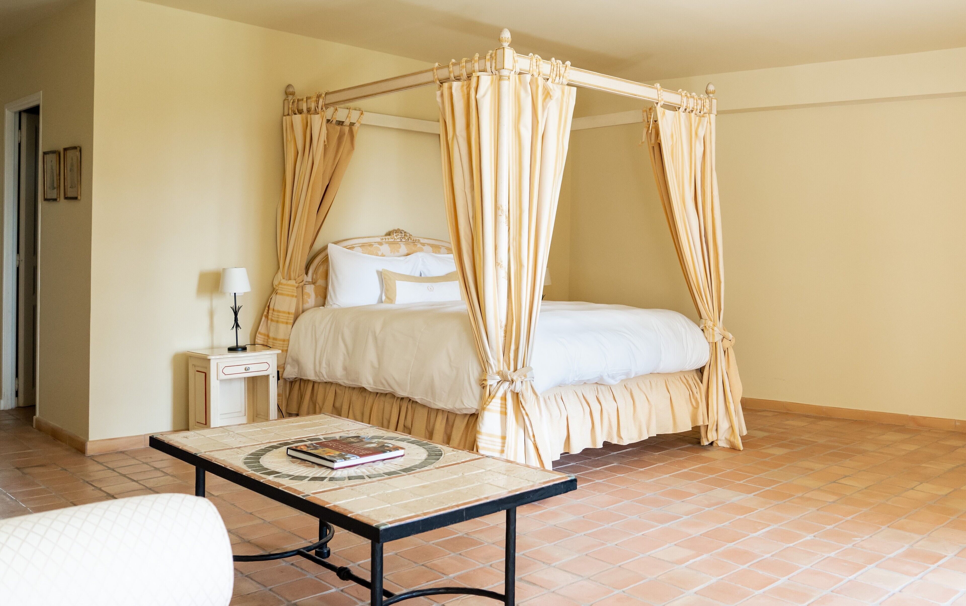 A bedroom with a four-poster canopy bed, a bedside table with a lamp, and a tiled floor.