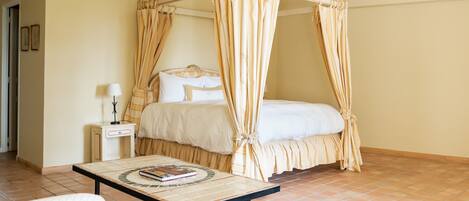 A bedroom with a four-poster canopy bed, a bedside table with a lamp, and a tiled floor.