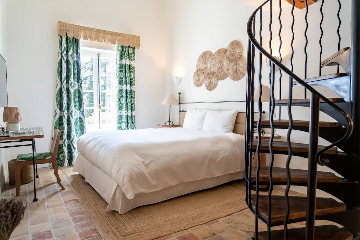 A bedroom with a bed, a desk, a lamp, a window with curtains, and a spiral staircase.