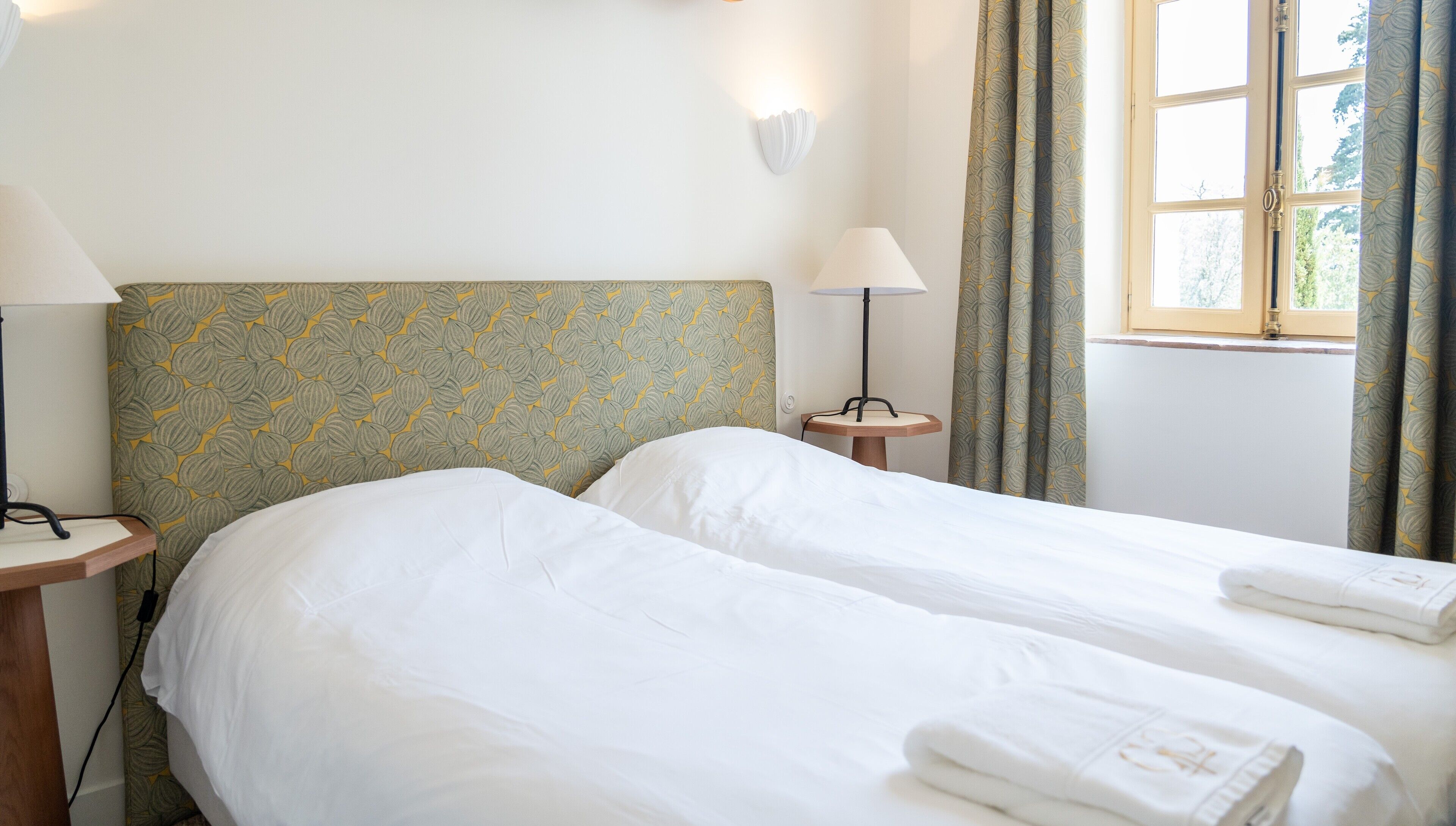Une chambre d’hôtel avec deux lits simples, une tête de lit à motifs, des lampes de chevet et une fenêtre avec des rideaux.