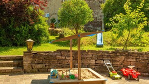 A sandbox with toys and a small tree, surrounded by a stone wall and greenery. - Rustic Eifel - Garden, kitchen - Hiking, bike trails & self-check-in (Gerolstein)