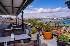 Un espace extérieur aménagé avec des tables et des chaises, des plantes en pot, et une vue sur la plage et le paysage urbain.