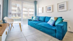Een moderne woonkamer met een blauwe bank, een witte fauteuil en een eettafel. Drie ingelijste foto’s hangen aan de muur.