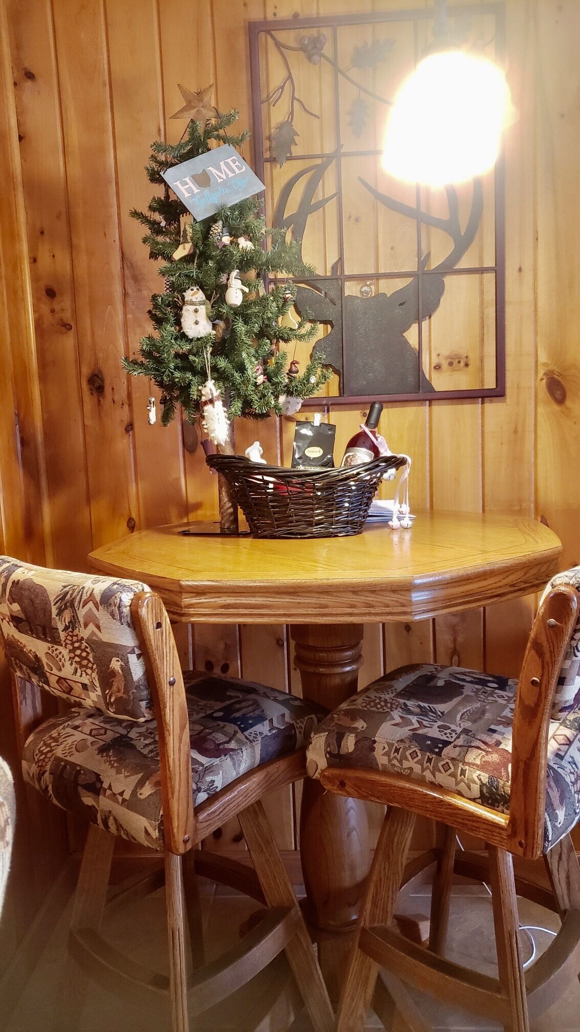 Un interior de madera con una mesa redonda, dos sillas estampadas, un árbol de Navidad y un adorno en la pared.