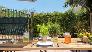 Un área de comedor al aire libre con una mesa dispuesta para dos personas, una valla metálica y diversa vegetación en el fondo.