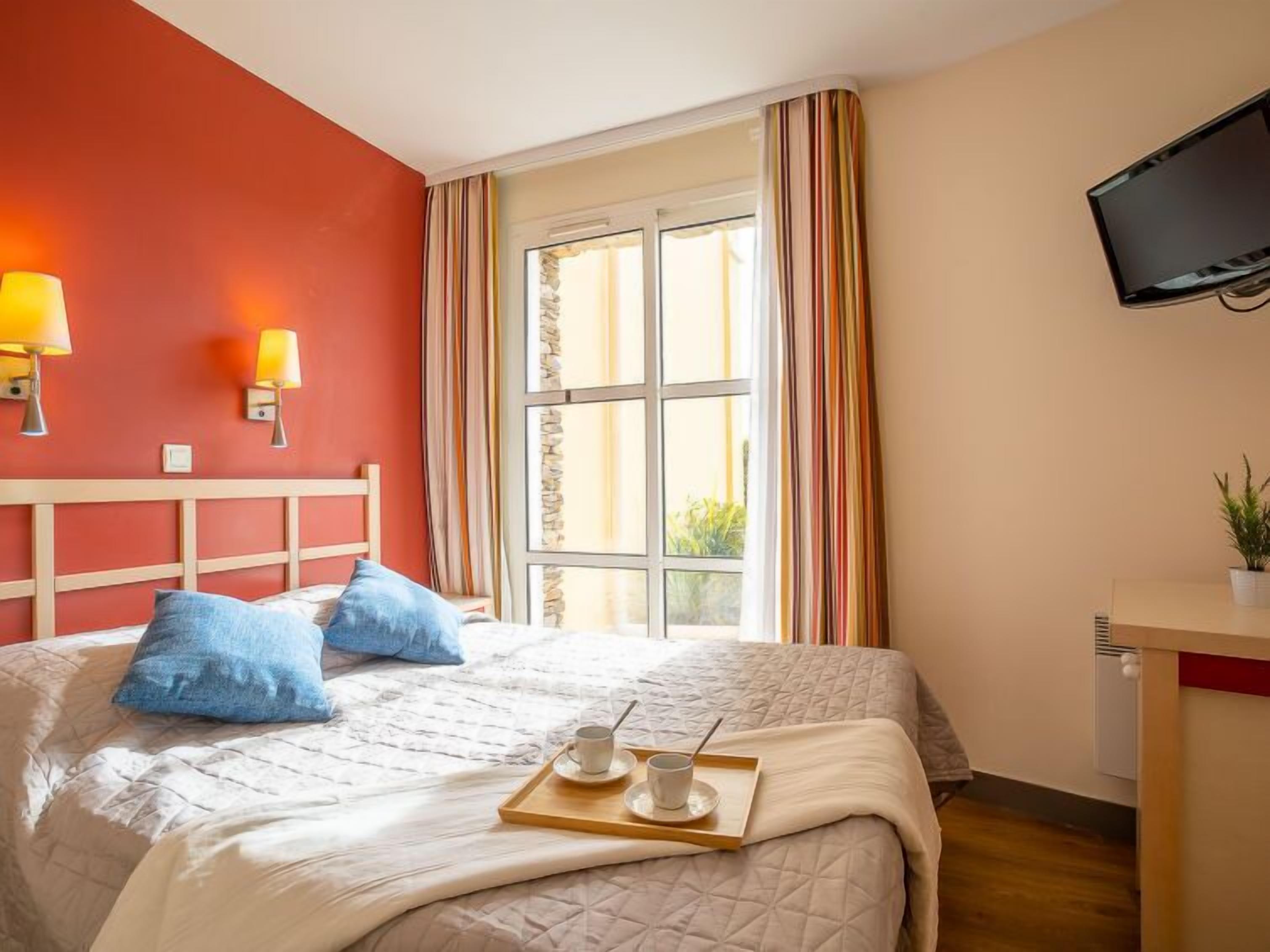 A hotel room with a bed, a TV, a window with curtains, and a tray with a cup and saucer.