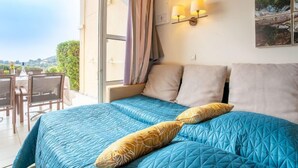 A bedroom with a bed, two pillows, a nightstand, a window with curtains, and a view of the outdoors. - Le Village (C4 008) by Interhome (Agay Village Cap Esterel)