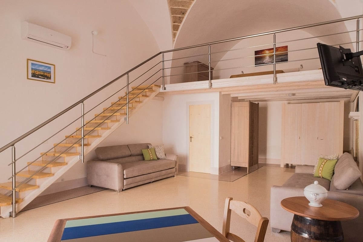Amplia sala de estar con escalera, sofá, mesa de centro y televisor montado en la pared.