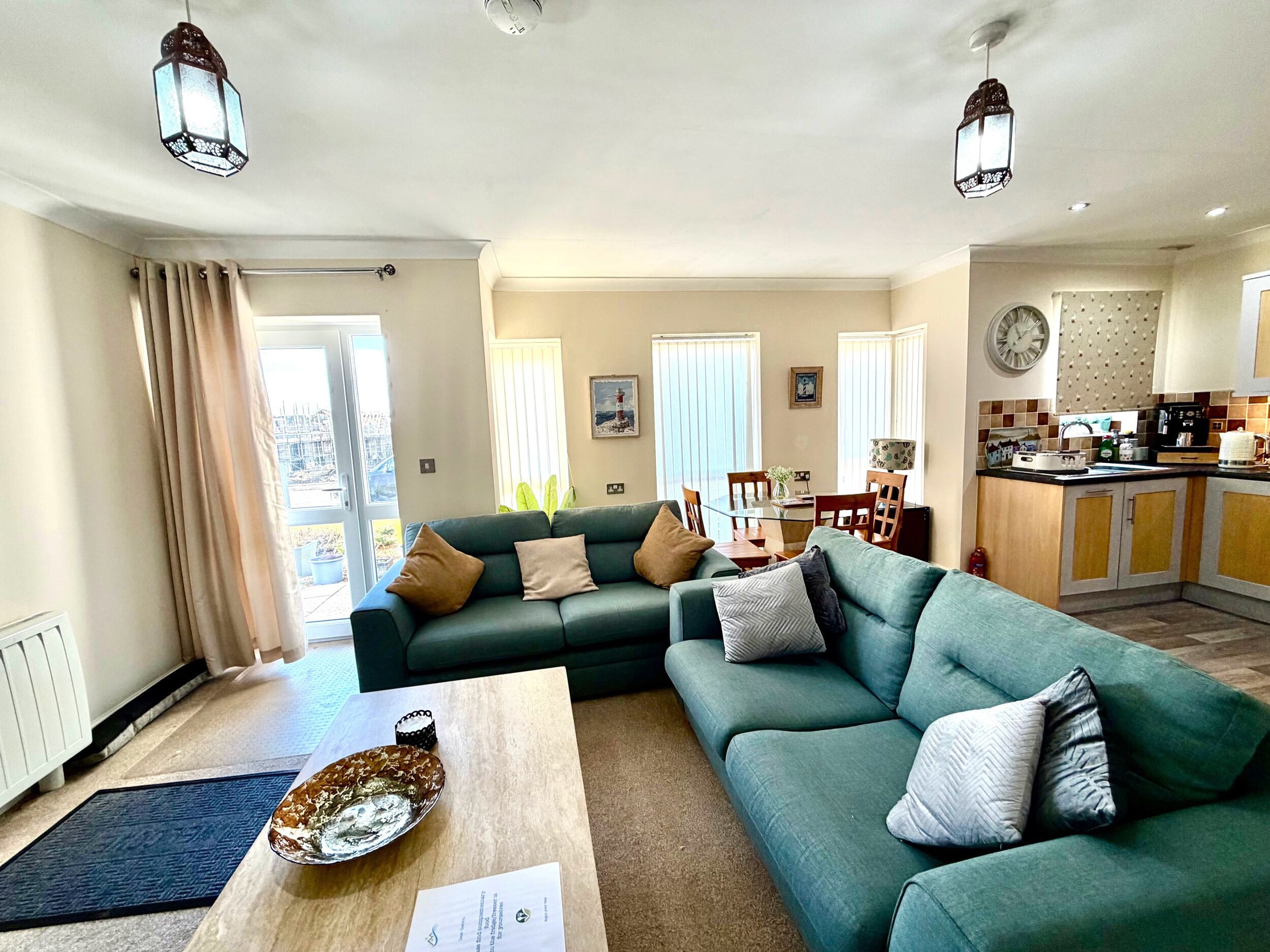 A living room with a sofa, coffee table, and a dining area with chairs and a table. There is a kitchen area with cabinets and a clock on the wall.