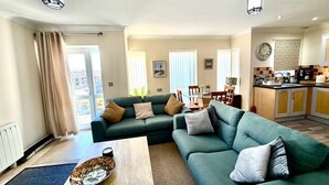 A living room with a sofa, coffee table, and a dining area with chairs and a table. There is a kitchen area with cabinets and a clock on the wall. - Apartment 1 (Morecambe)