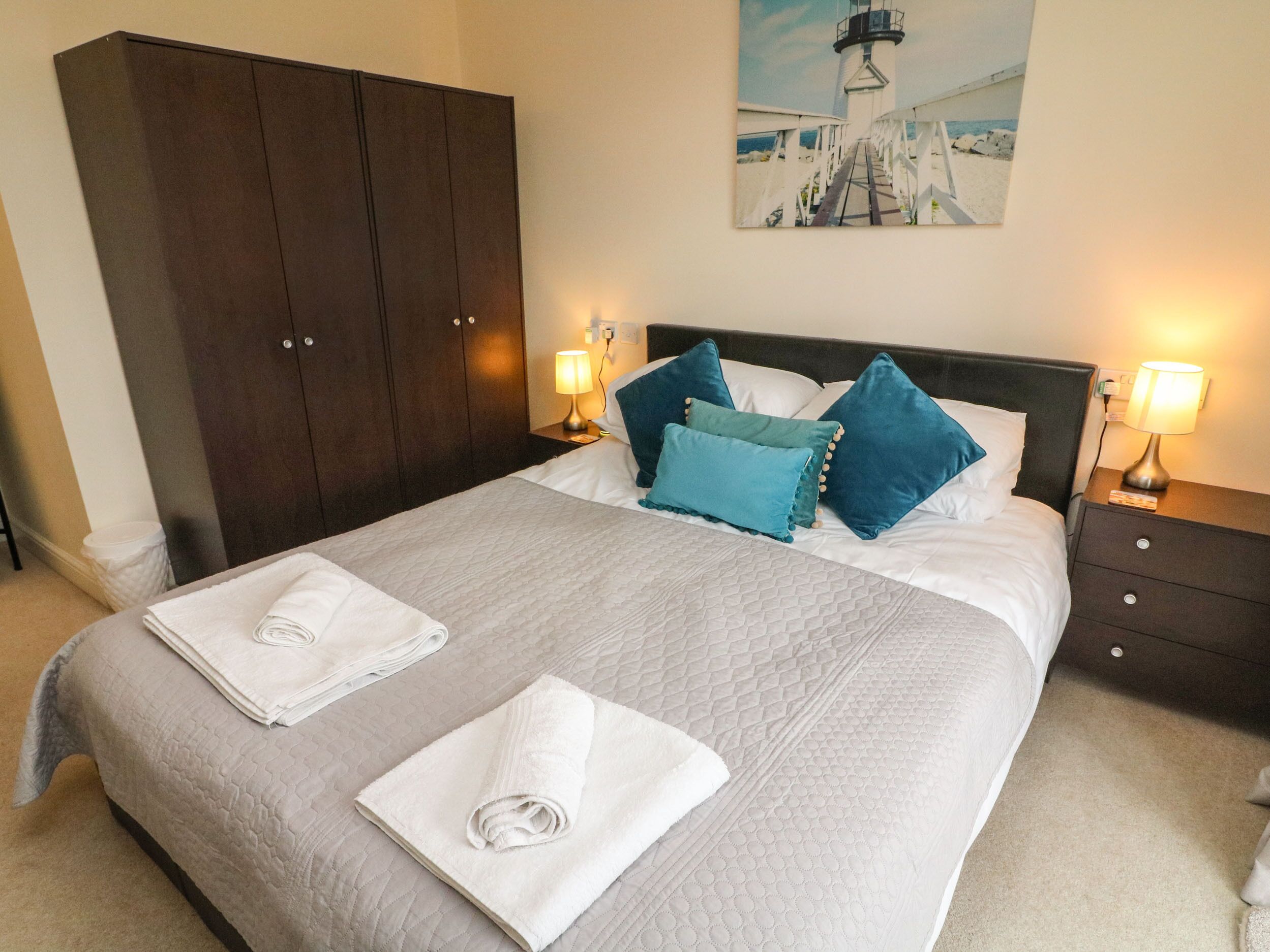 Una cama bien hecha con colcha gris acolchada, dos toallas blancas enrolladas y almohadas azules. A la izquierda, un armario de madera oscura, y en la pared, un cuadro de un faro.
