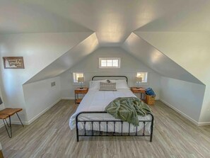 Une chambre avec un plafond en pente, un lit recouvert de draps blancs, deux lampes de chevet, une petite chaise en bois et une fenêtre.