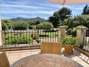 A patio with a tiled table and chairs, surrounded by a metal fence, overlooking a landscaped garden and distant hills.
