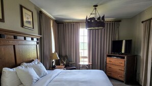 Ein Hotelzimmer mit einem großen Bett, einem Schreibtisch mit Lampe, einem Sessel, einem Fernseher auf einer Holzkommode und einem Fenster mit Vorhängen.