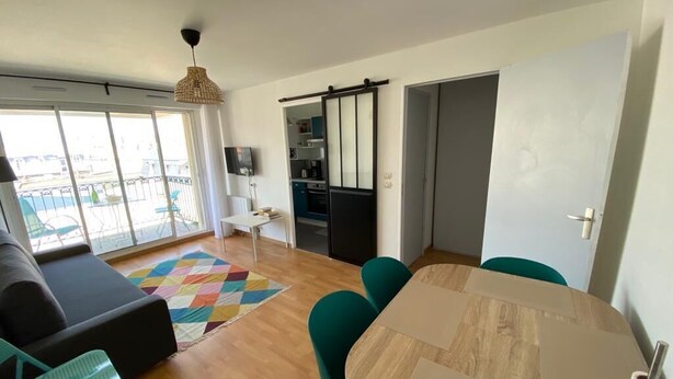 Una sala de estar moderna con un sofá, una mesa de comedor y sillas. Se observa un área de cocina al fondo con armarios y una encimera.