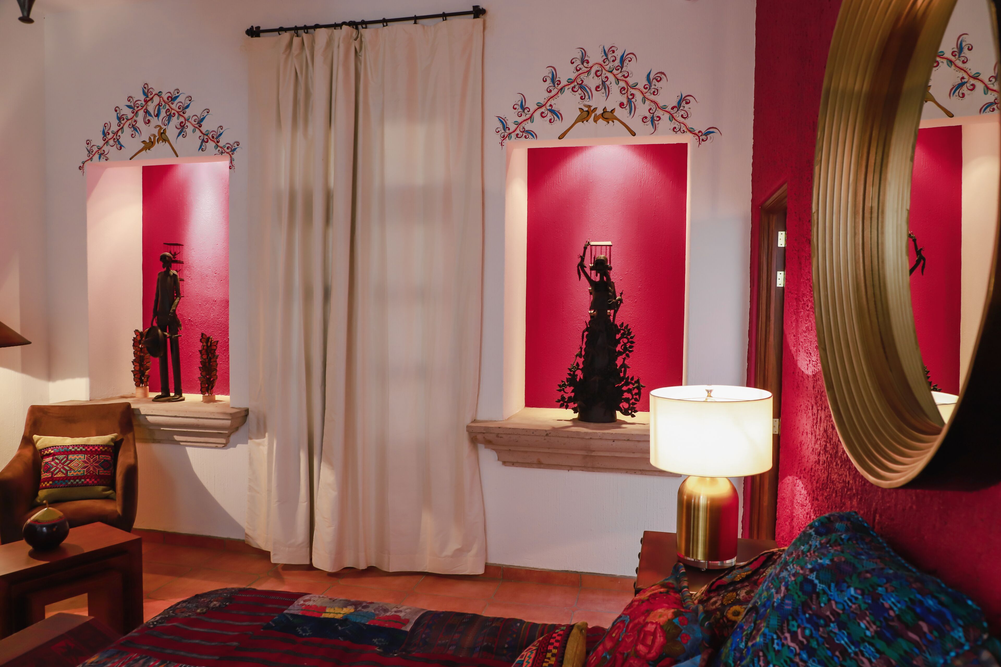 A room with a bed, a chair, a lamp, a mirror, and two wall-mounted sculptures.