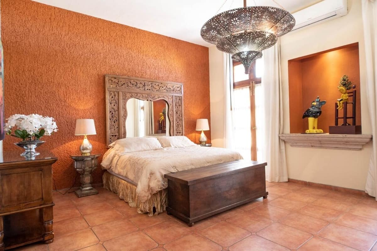 Ein Schlafzimmer mit einem großen Bett, zwei Nachttischen inklusive Lampen, einer hölzernen Bank, einem dekorativen Spiegel und einer Kristallleuchtre.