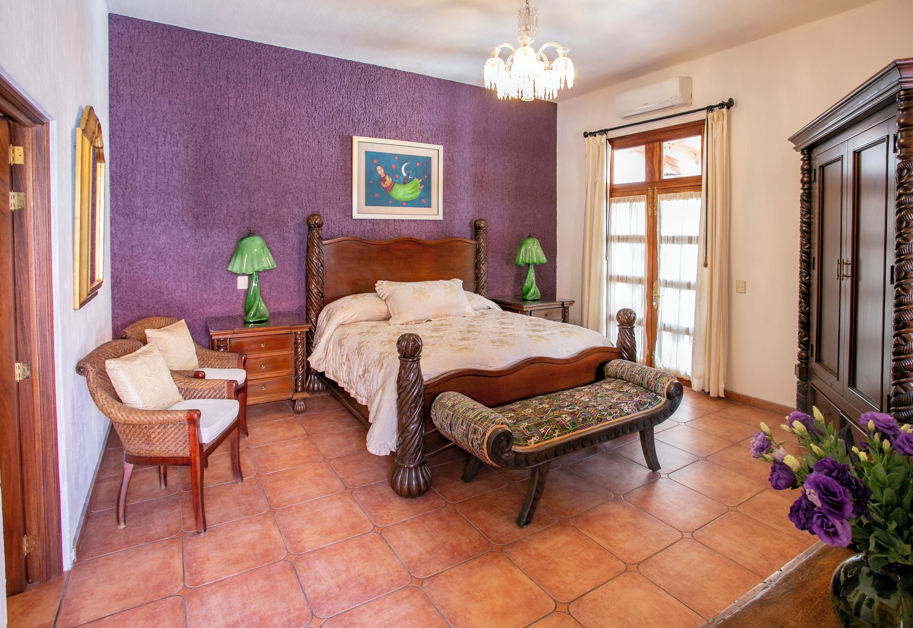 Quarto com uma cama grande, um banco de madeira, duas mesas de cabeceira, um abajur verde e uma parede roxa.