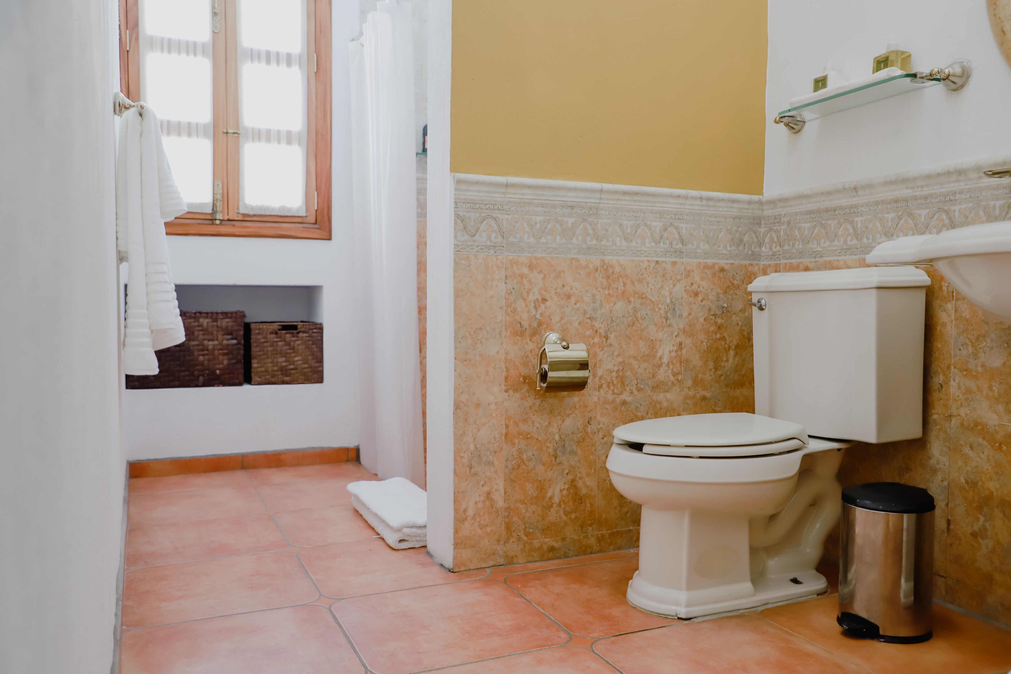 A bathroom with a toilet, a trash can, a towel rack, and a shelf with toiletries.