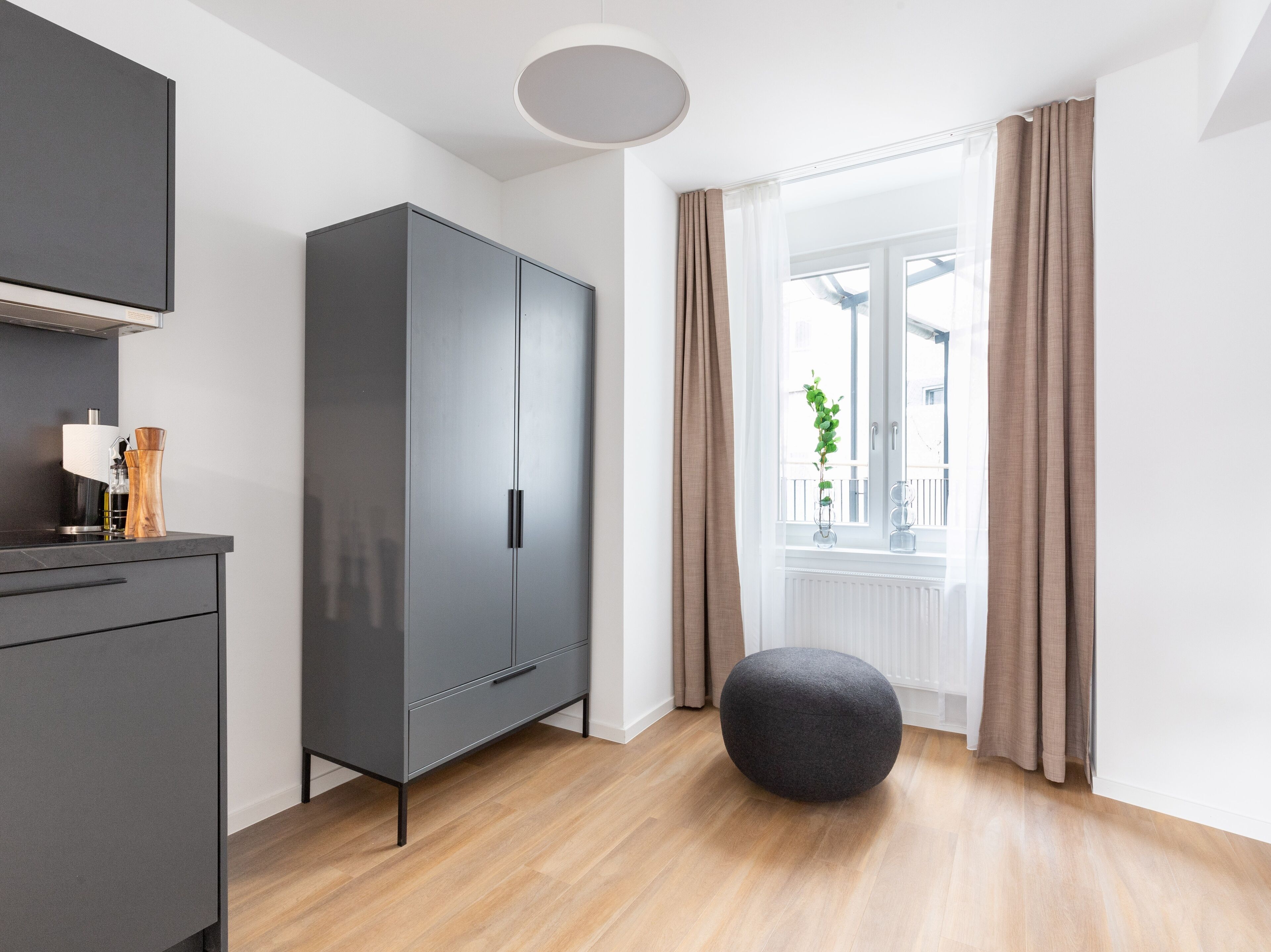 A modern room with a grey wardrobe, a round ottoman, and a window with curtains.